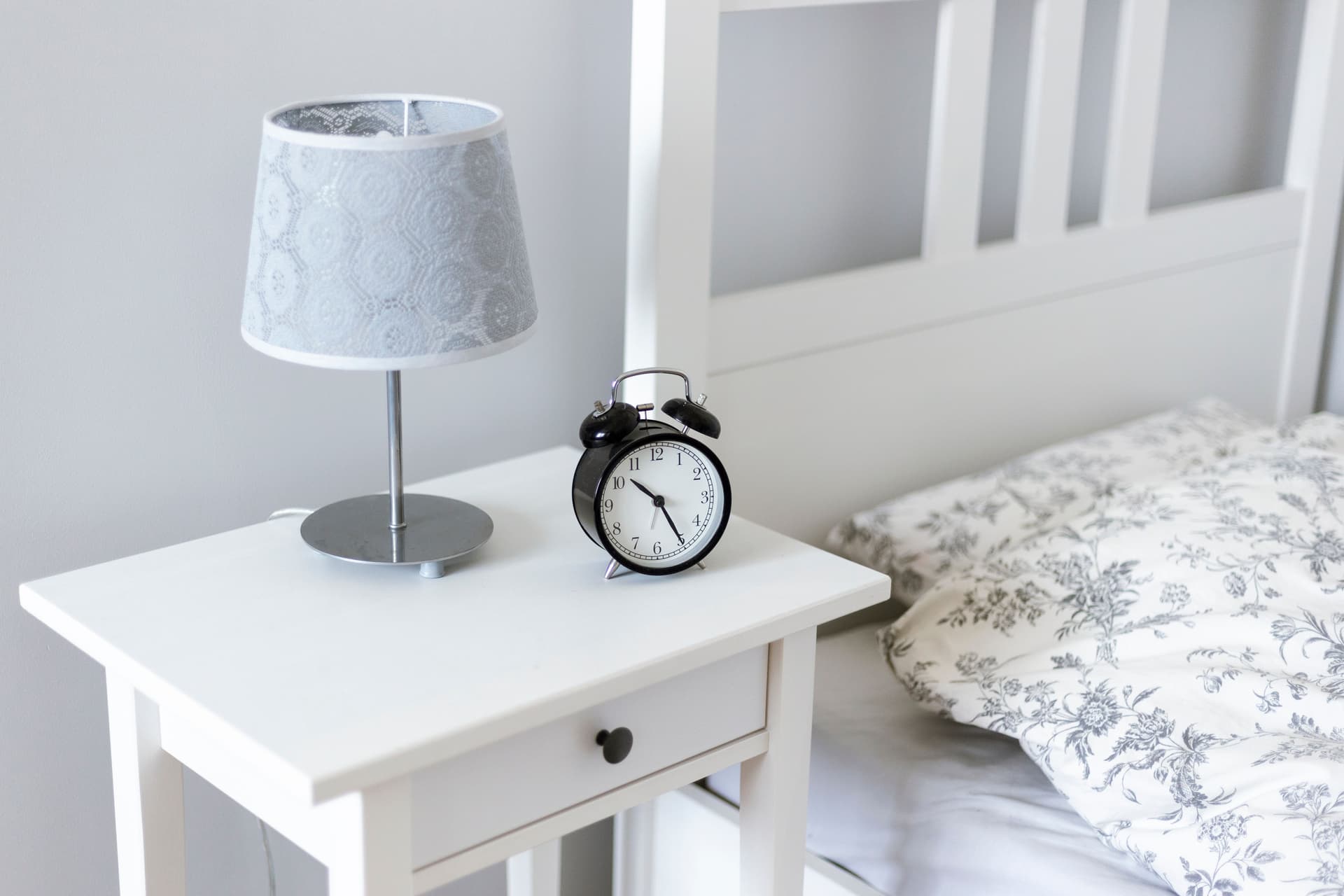 A white bedside table with an alarm clock and a lamp beside a bed
